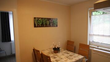 A dining area with a table set for two, a potted plant, and a wall-mounted painting of plants.
