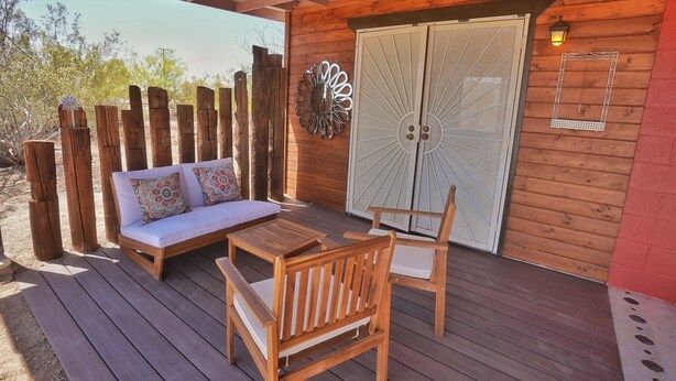 A wooden deck with a sofa and two chairs, a decorative wall piece, and a double door. - Groovy Desert Getaway w Pool, Fire pit, Stream/Wifi, Vinyl Records, Books, Art.. (Twentynine Palms)