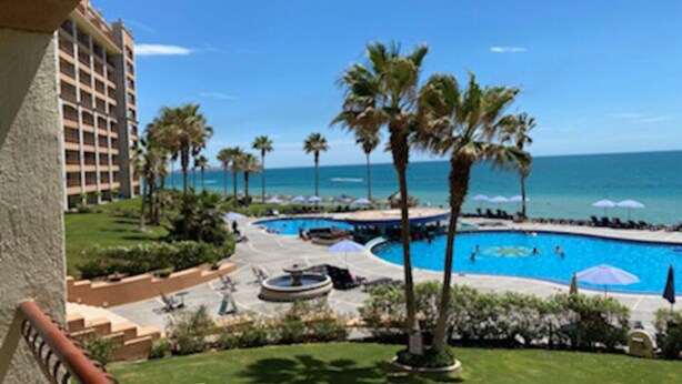 Ein Poolbereich eines Hotels mit Palmen, blauem Himmel und Blick aufs Meer.
