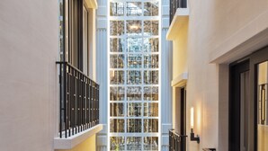 A narrow indoor corridor with a glass wall on one side, allowing a view of a garden with plants and a table with chairs. - The Alest Hotel (Mexico City)