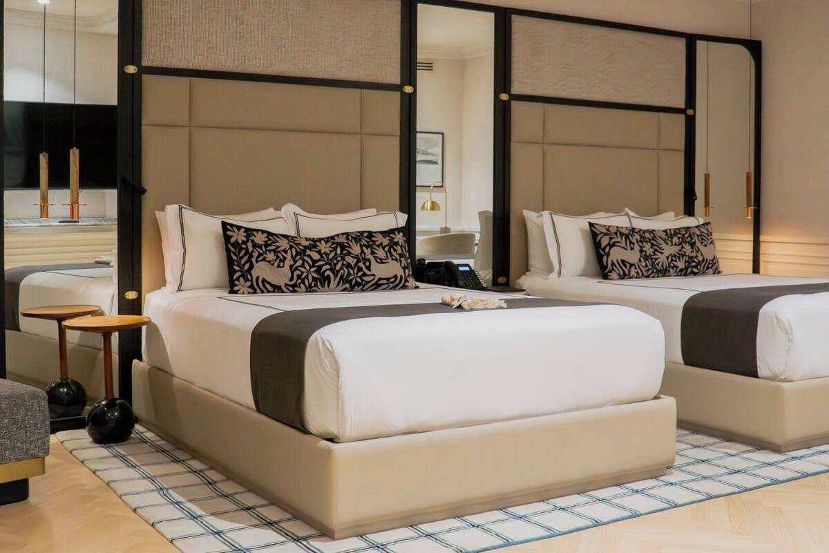 A modern hotel room with two beds, a large mirror, and a small table with a lamp.