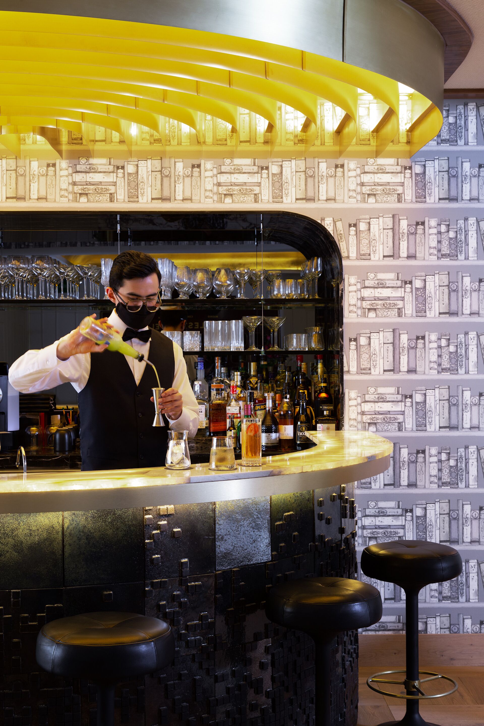 Uma área de bar com um bartender preparando uma bebida, prateleiras com garrafas, copos e uma parede estampada.