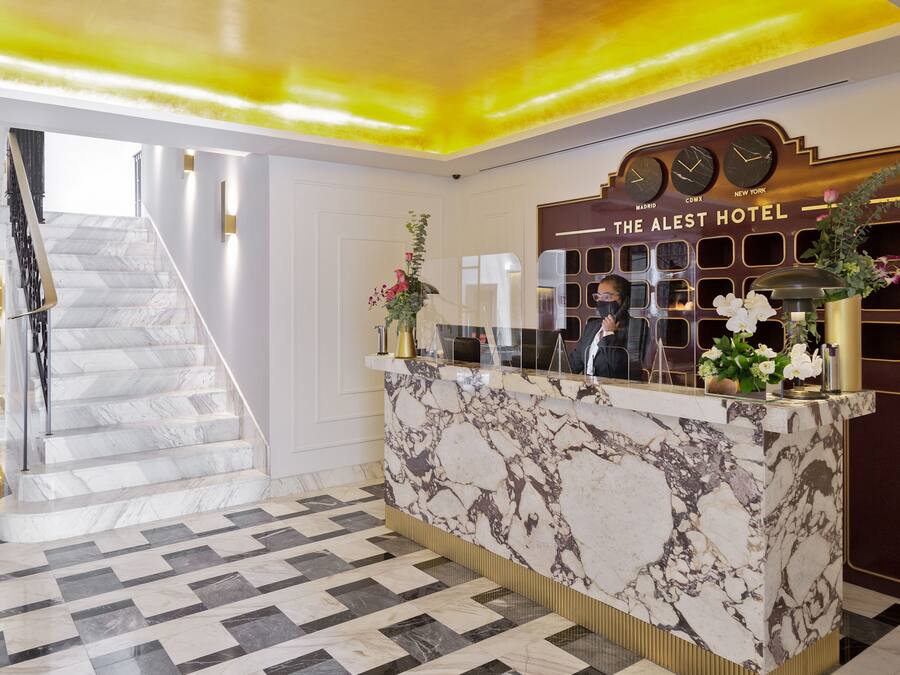 Ein Hotel-Empfangsbereich mit einer Marmor-Theke, einer Uhr, die die Zeit in drei Städten anzeigt, und einer Treppe zu einer anderen Etage.