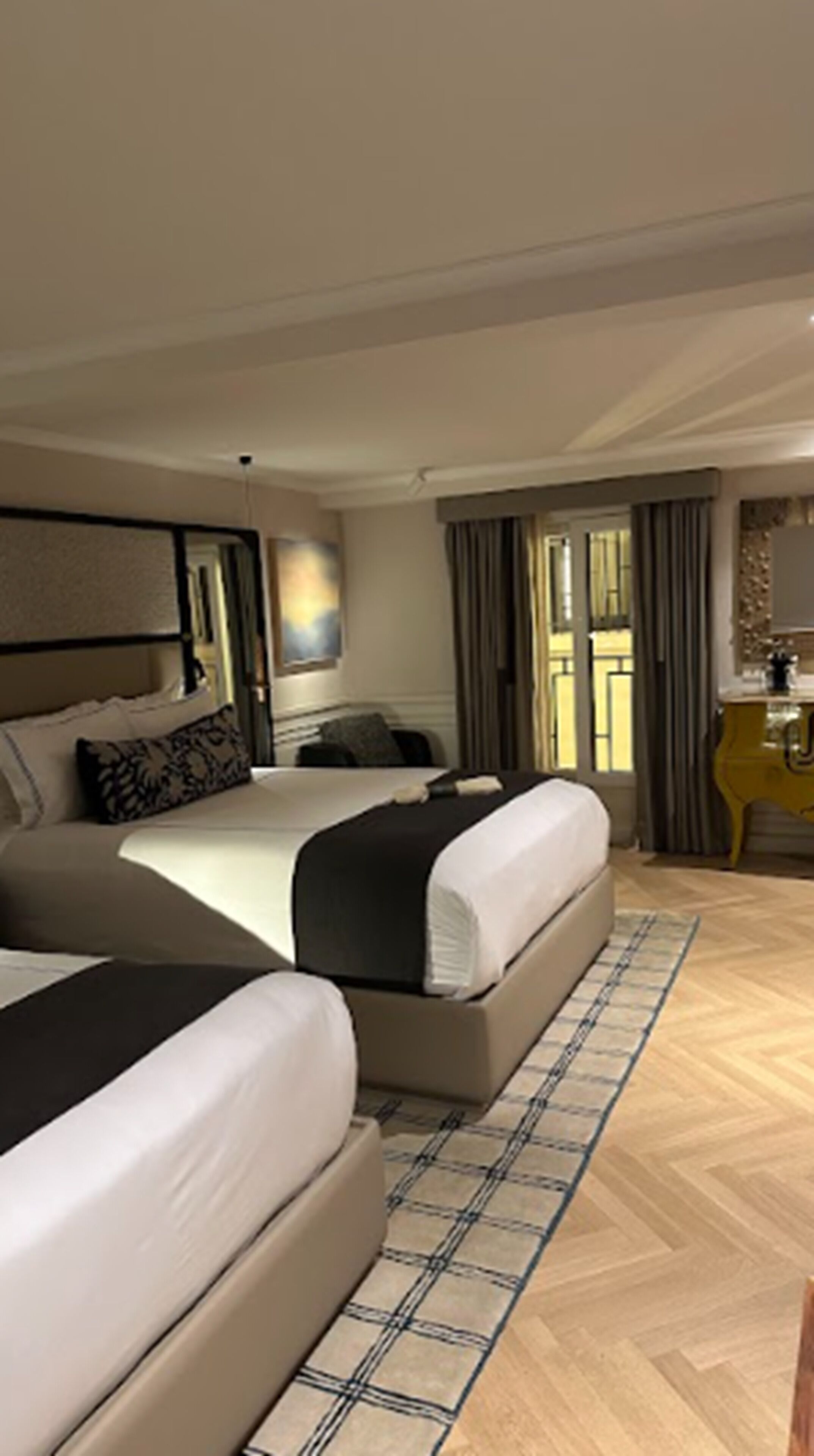 A hotel room with two beds, a large window with curtains, a wooden floor, and a small desk with a chair.