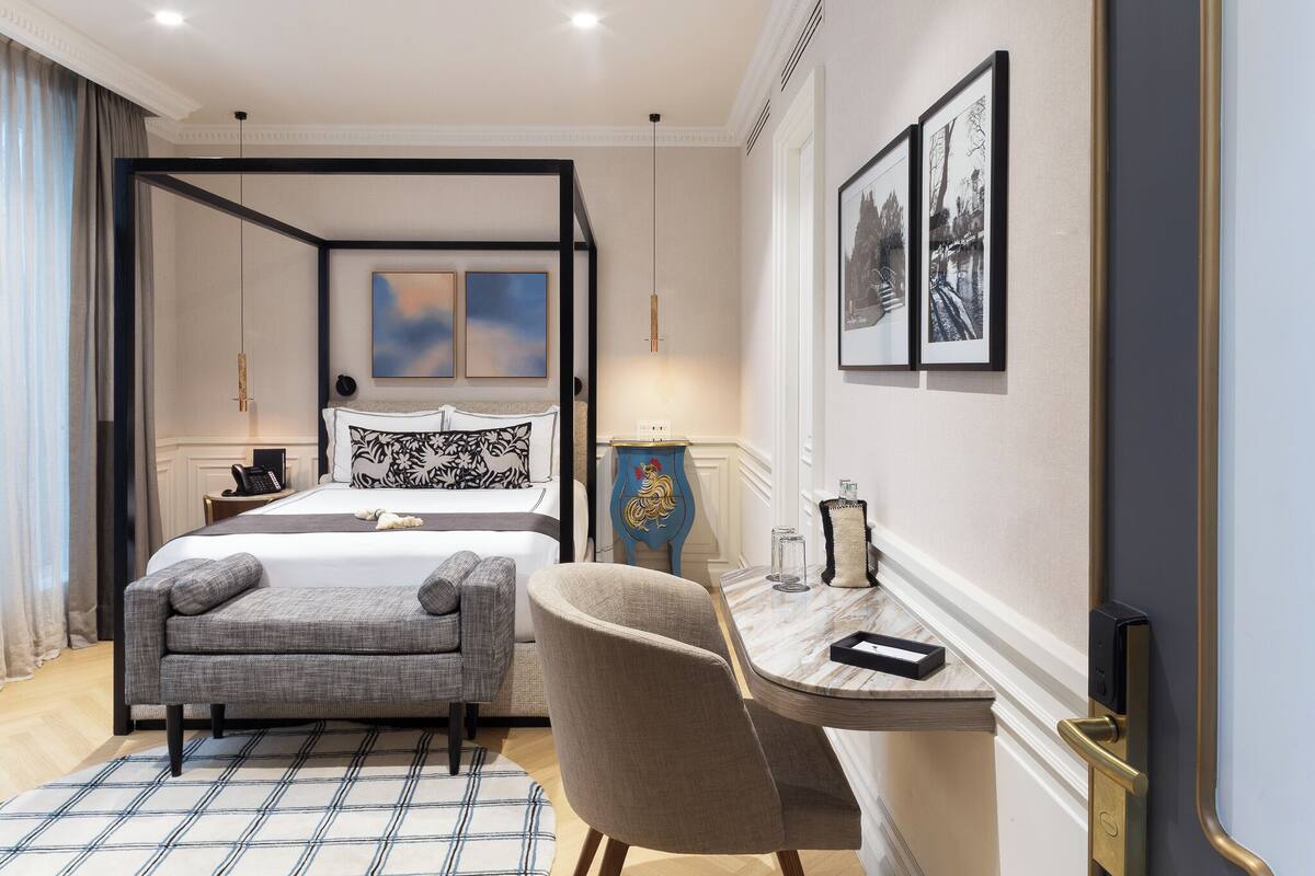 A modern hotel room with a four-poster bed, a desk with a chair, and a view of the city.