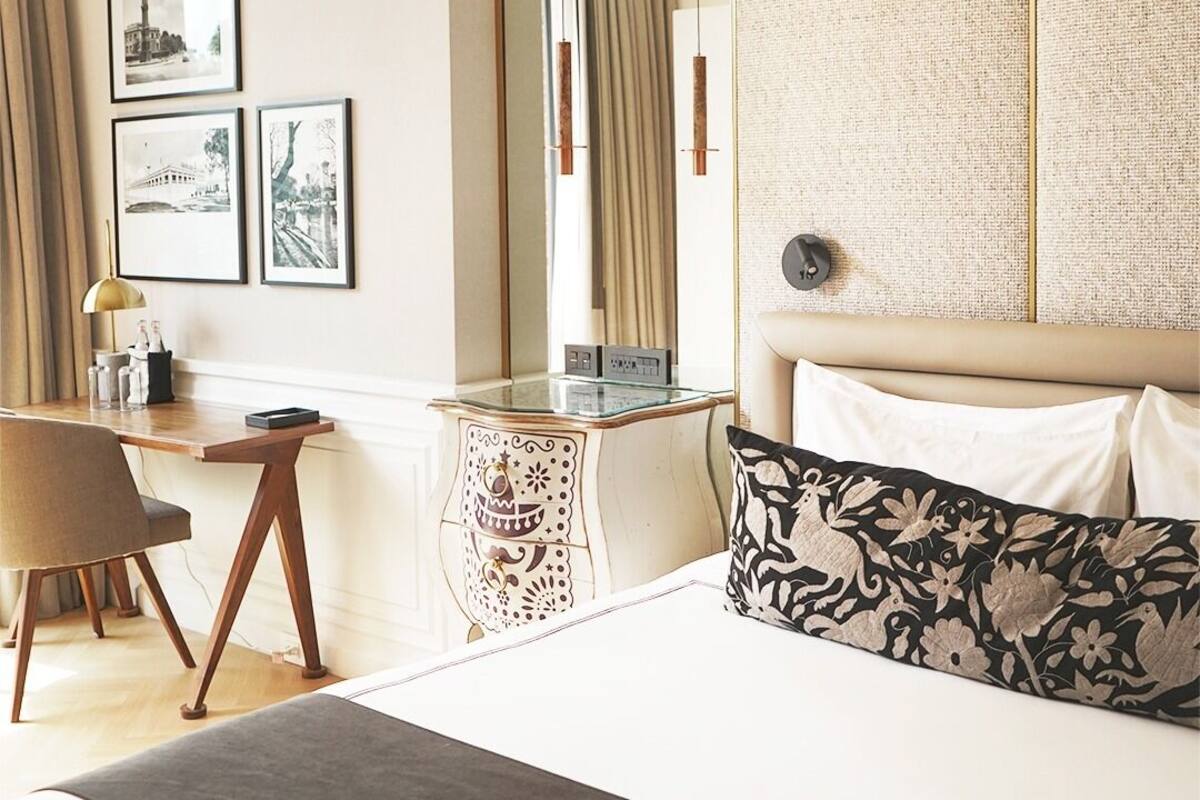 A hotel room with a bed, a desk, and a chair. There are framed pictures on the wall and a lamp on the desk.