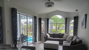 A spacious living room with a grey sofa, a patterned ottoman, and a marble table. Large windows offer views of a swimming pool and greenery. - 4 Bedroom Pool home with pond view in Sarasota, FL (Sarasota)