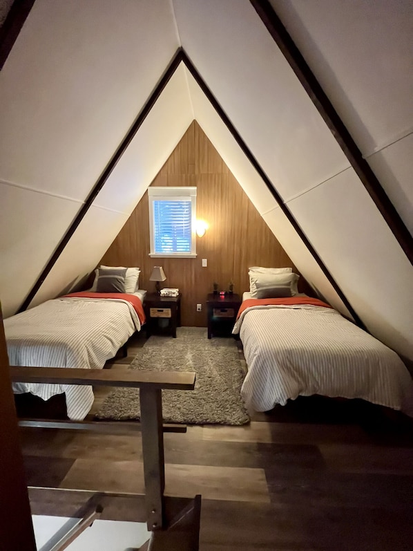 A triangular attic bedroom with two beds, a small window with blinds, and a nightstand with a lamp.