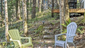 Two Adirondack chairs in a forest setting with a stone fire pit and a wooden deck in the background. - Timberline cabin close to Mt. Rainer and White Pass, WiFi, Netflix, AC (Packwood)