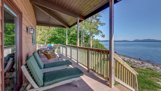 A wooden deck with two green lounge chairs and a table, overlooking a lake with a rocky shoreline. - Wings Cottage - Oceanfront! (Trenton)