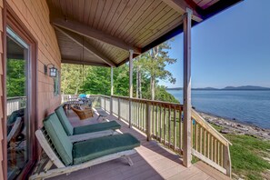 A wooden deck with two green lounge chairs and a table, overlooking a lake with a rocky shoreline. - Wings Cottage - Oceanfront! (Trenton)