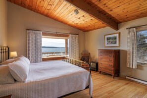A bedroom with a wooden ceiling, a bed with a quilted blanket, a dresser, a framed picture, and a window with curtains.