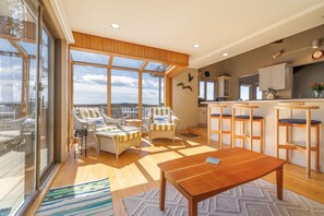 Relax in a spacious living area with a large table and comfortable seating, featuring a large window with an outdoor view and a modern kitchen area.