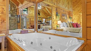 A wooden cabin interior with a large bathtub, a dining area, and a cozy living room with a couch and pillows. - Location! Cute Studio Log Cabin,Hot Tub,Fireplace (Pigeon Forge)