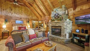 A cozy log cabin interior with a stone fireplace, a comfortable sofa, and a television mounted on the wall. - Location! Cute Studio Log Cabin,Hot Tub,Fireplace (Pigeon Forge)