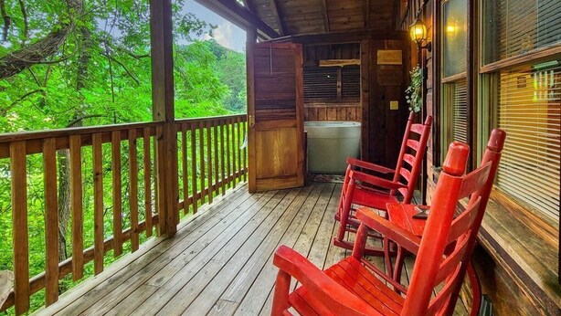 A wooden porch with red chairs, a ceiling fan, and a view of lush greenery. - Location! Cute Studio Log Cabin,Hot Tub,Fireplace (Pigeon Forge)