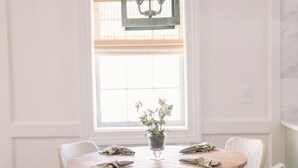 Un coin repas avec une table ronde en bois entourée de six chaises en osier, un luminaire de style lanterne vert au-dessus, et une fenêtre avec des stores.