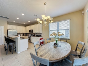 A modern kitchen with white cabinets, a central island, and a dining area with a round wooden table and chairs. - Veranda Home, 8br, 6 en-suites, Lake View, POOL/SPA, FREE Resort Amenities! (Kissimmee)