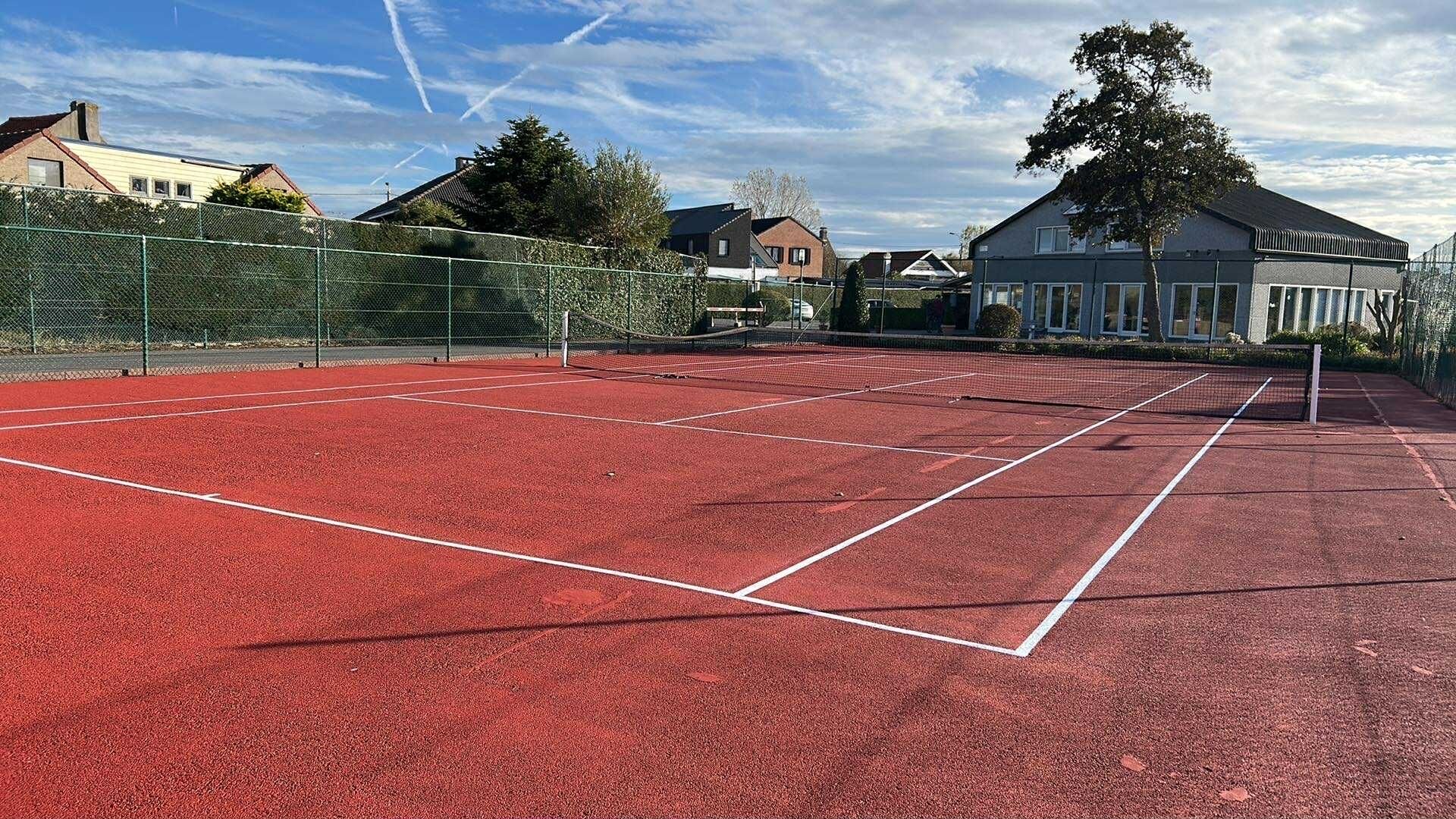 Un court de tennis à surface rouge, entouré d’une clôture en treillis métallique, avec des maisons en arrière-plan.