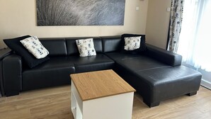 A modern living room with a black leather sofa, two patterned pillows, a wooden coffee table, and a large landscape painting on the wall. - Family apartment for 6 people (De Haan)