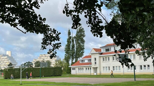 A park with a basketball court, a large building with a red roof, and a hedge. - Family apartment for 6 people (De Haan)