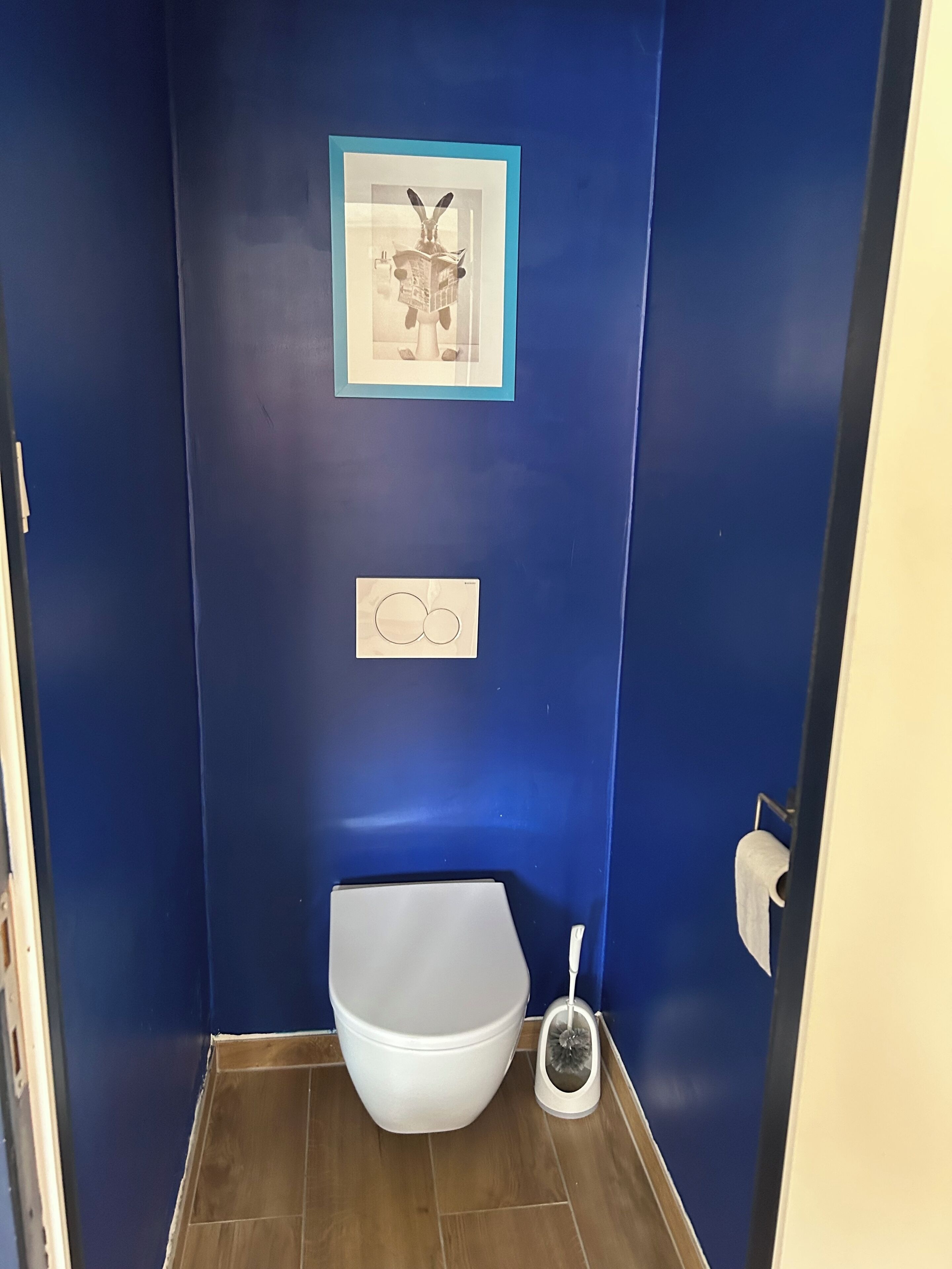 Une petite salle de bain avec un mur bleu, des toilettes blanches et un tableau encadré au-dessus des toilettes.