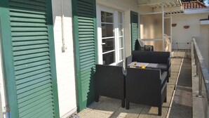 A covered outdoor seating area with wicker furniture and a table, adjacent to a building with green shutters and a white exterior. - Family apartment for 6 people (De Haan)