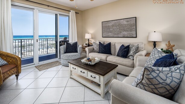 Amplia sala de estar con un gran ventanal corredero que da a un balcón con vista a la playa. Hay un sofá color beige, un sillón de mimbre y una mesa de centro con objetos decorativos.