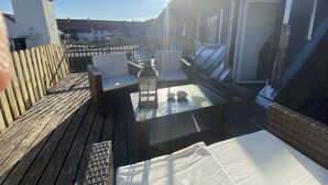 Terraço no telhado com móveis de vime, uma mesa de vidro e vista para os edifícios ao redor.