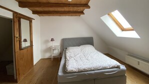 Quarto com teto inclinado, claraboia, cama, mesa de cabeceira, abajur e porta.