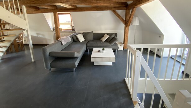 Um loft moderno com vigas de madeira aparentes, um sofá modular cinza, uma mesa de centro branca e uma escada com corrimão branco.