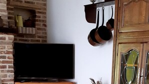 Een eethoek met een ronde tafel met een tafelkleed met een patroon, houten stoelen, een bakstenen muur, een televisie op een houten dressoir, en een kast met hangende potten.