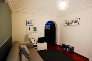 A room with a sofa, a small table with a lamp, a TV, and a bookshelf. There is a framed picture on the wall and an archway leading to another room.