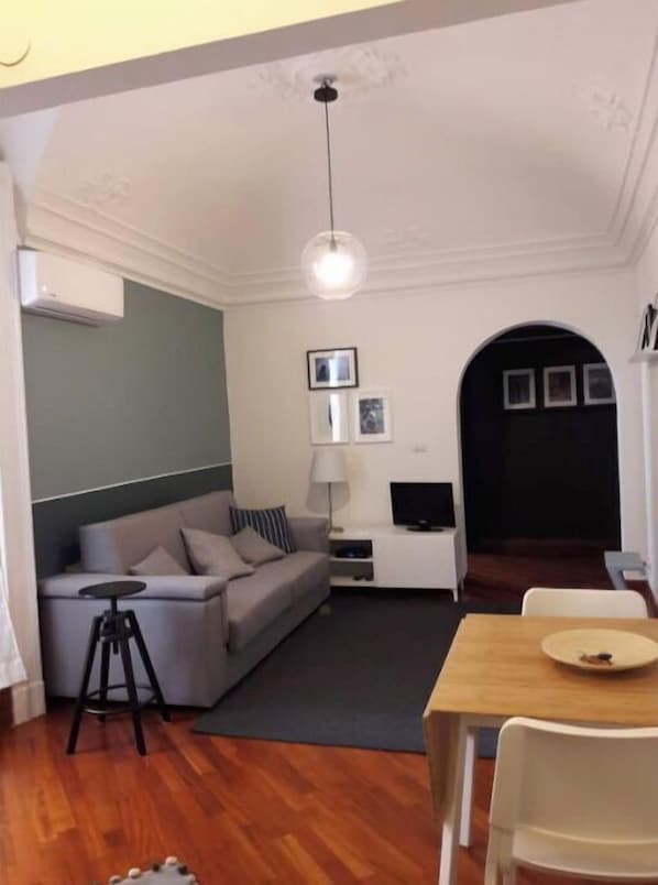 A modern living room with a sofa, a small table, a TV, and a dining area with chairs.