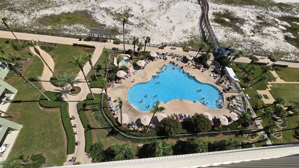 An aerial view of a resort with a central swimming pool, surrounded by palm trees and a sandy beach area.