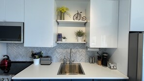 A modern kitchen with a stainless steel sink, a kettle, and a toaster. There are shelves with decorative items and a herringbone-patterned backsplash. - Sage View Suites (Peachland)
