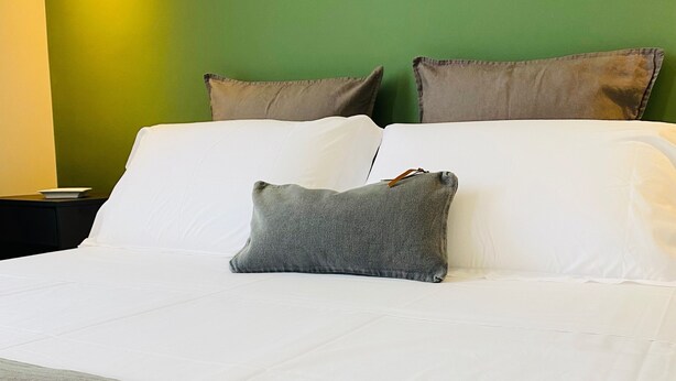 Een netjes opgemaakt bed met witte lakens en een grijs kussen, een groene muur en een nachtkastje met een wit bord.