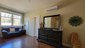 A spacious room with hardwood flooring, a dark-colored sofa with a patterned pillow, a large mirror above a dark wooden dresser, and a wall-mounted air conditioner. - Spacious apartment, deep in the woods, with private entrance and deck (Cleveland)
