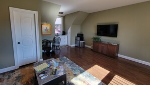 A living room with a dining area, a kitchen, and a TV mounted on the wall. - Spacious apartment, deep in the woods, with private entrance and deck (Cleveland)
