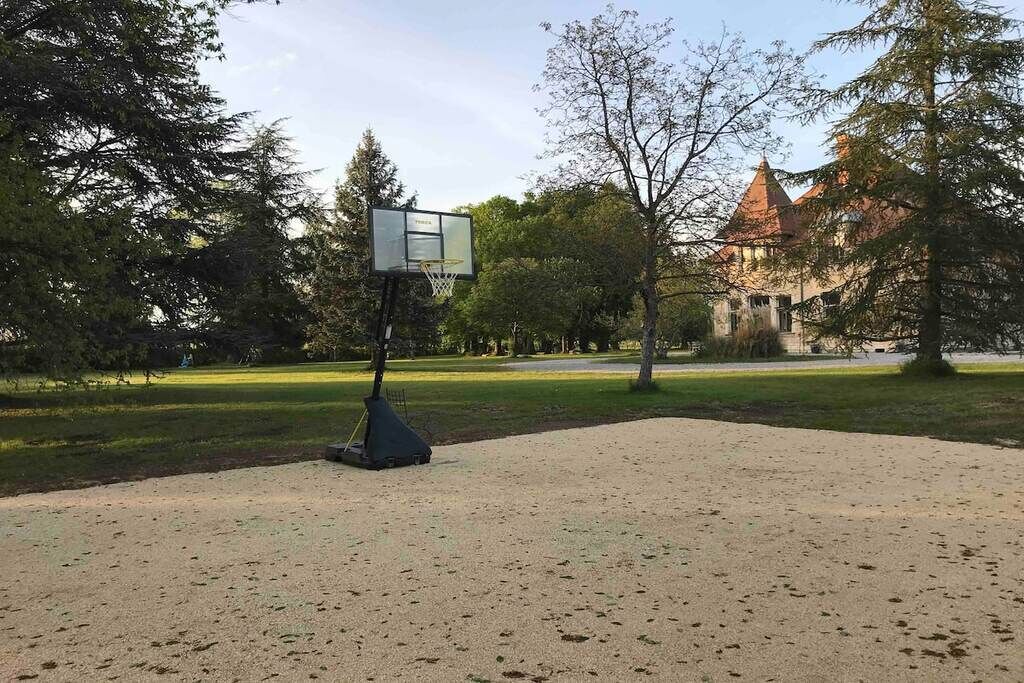Ein Basketballplatz mit Korb, umgeben von Bäumen und einem Gebäude mit rotem Dach im Hintergrund.