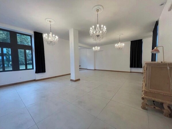 A spacious room with large windows, a chandelier, and a wooden cabinet.