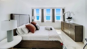 Une chambre à coucher comprenant un lit, des tables de chevet, une commode, une fenêtre avec des rideaux et une vue sur la mer.