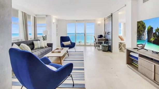 Un salon moderne avec un canapé, des fauteuils bleus, un téléviseur et une vue sur l’océan.