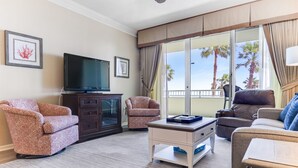 Una sala de estar con una televisión de pantalla plana, dos sillones estampados, un sofá, una mesa de centro y vistas a palmeras a través de grandes ventanales.