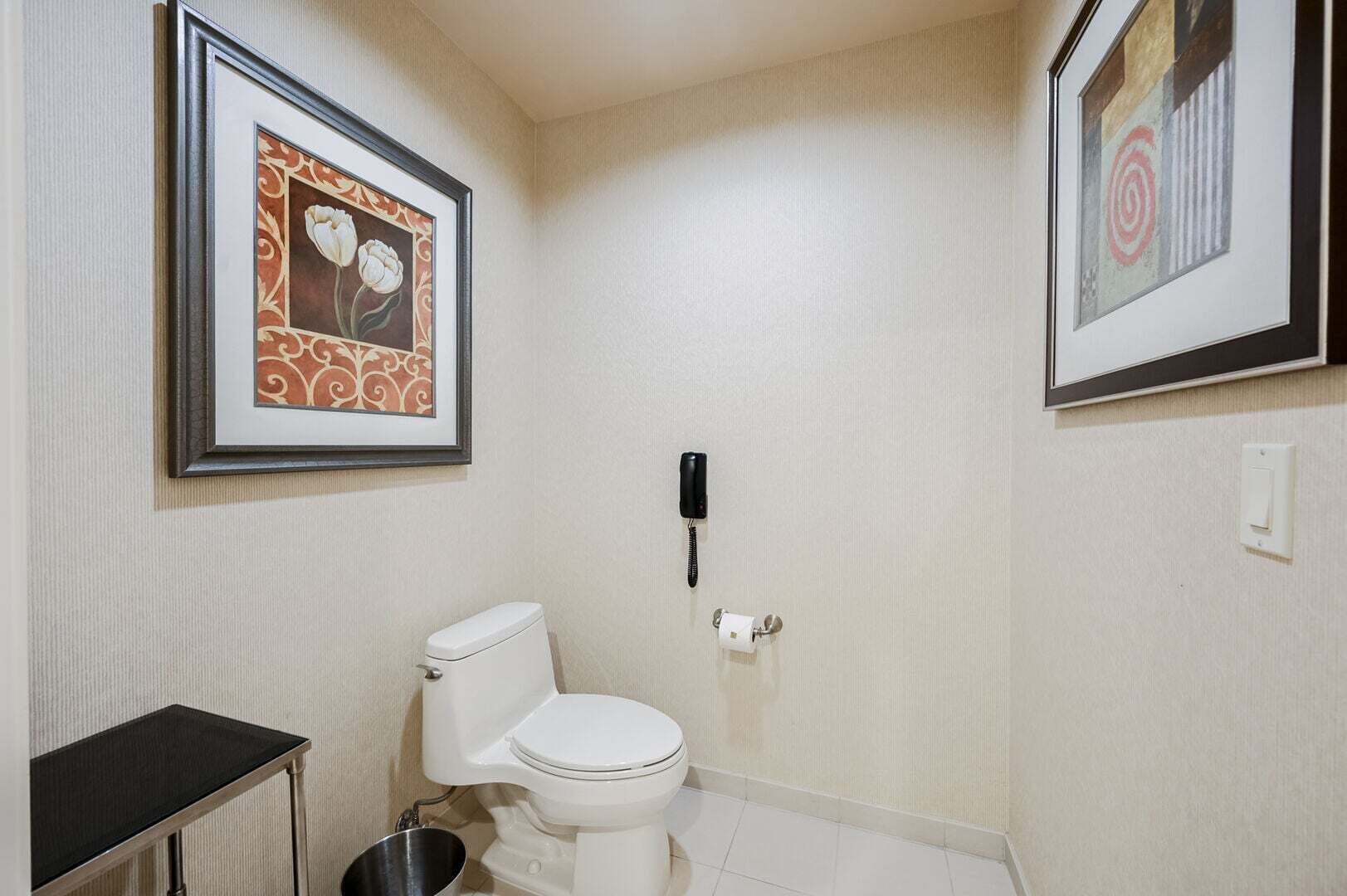 A small bathroom with a toilet, a trash bin, a framed picture on the wall, and a telephone.