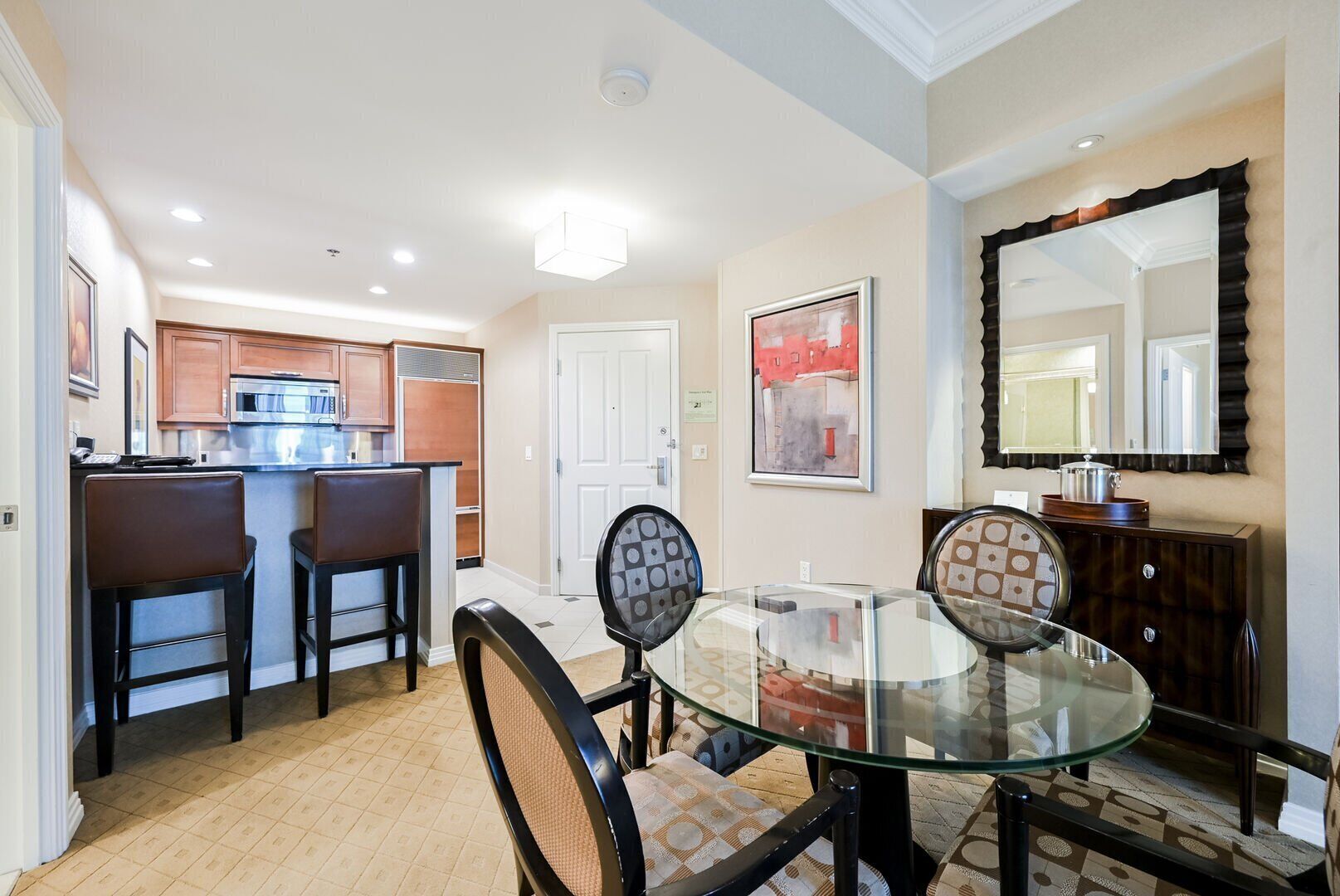 A dining area with a glass table and chairs, a kitchen in the background, and a large mirror on the wall.