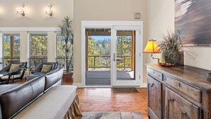 A spacious living area with a leather sofa, a wooden cabinet, and a balcony with a view of trees. - BEAUTIFUL HOME OVERLOOKING THE RIVER-MINUTES TO IT ALL-UPDATED KITCHEN! (Bend)