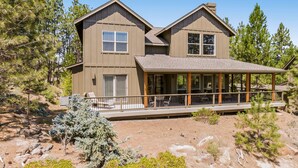A two-story house with a covered porch and a deck, surrounded by trees and rocky terrain. - BEAUTIFUL HOME OVERLOOKING THE RIVER-MINUTES TO IT ALL-UPDATED KITCHEN! (Bend)