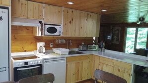 Une cuisine avec des meubles en bois, un four à micro-ondes, un grille-pain, une machine à café et un lave-vaisselle. On y trouve deux chaises en bois et une fenêtre donnant sur un paysage verdoyant.
