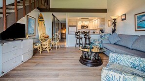 A spacious living area with a sofa, a coffee table, and a television on a cabinet. A staircase leads to an upper level. - Spacious 3bd Papakea condo, the perfect getaway (Lahaina)